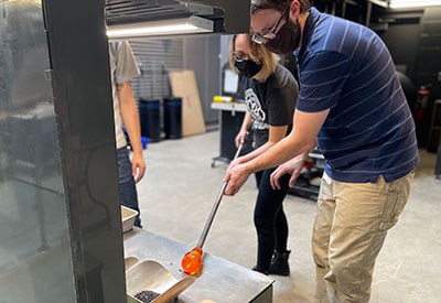 Two people standing working on shaping molten glass on a punti rod